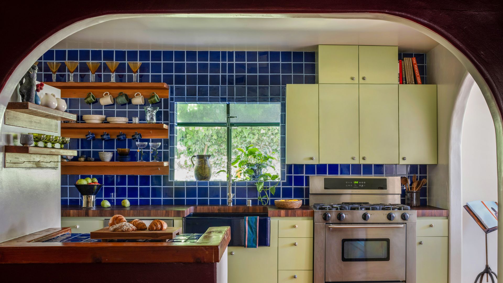 An arched ceiling leading into a kitchen with sage green cabinets, a stainless steel stove and oven, striking cobalt blue tiled backsplash, floating wood shelves adorned with glassware, and a wooden counter