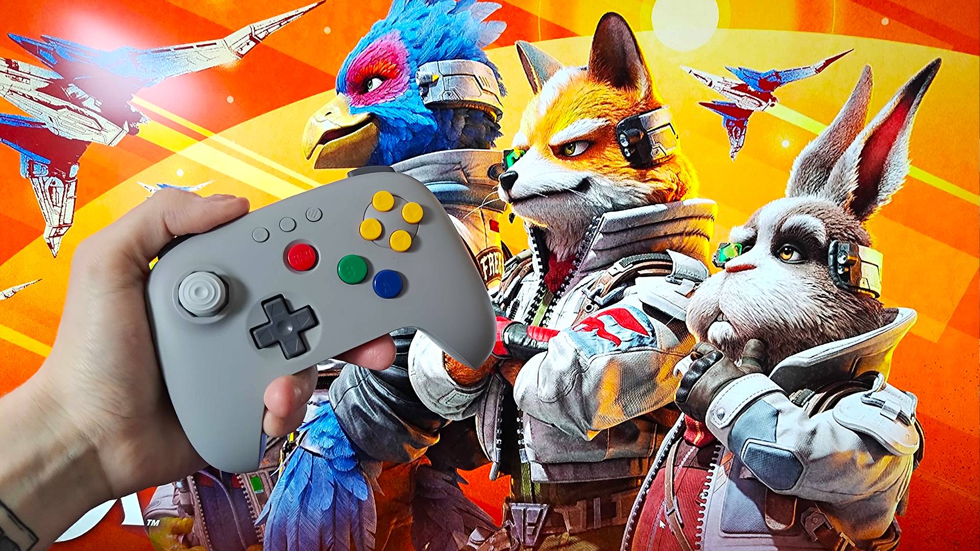 Hand holding gray 8Bitdo N64 controller in front of Star Fox for Switch 2 official artwork with Fox and Peppy looking to the left.