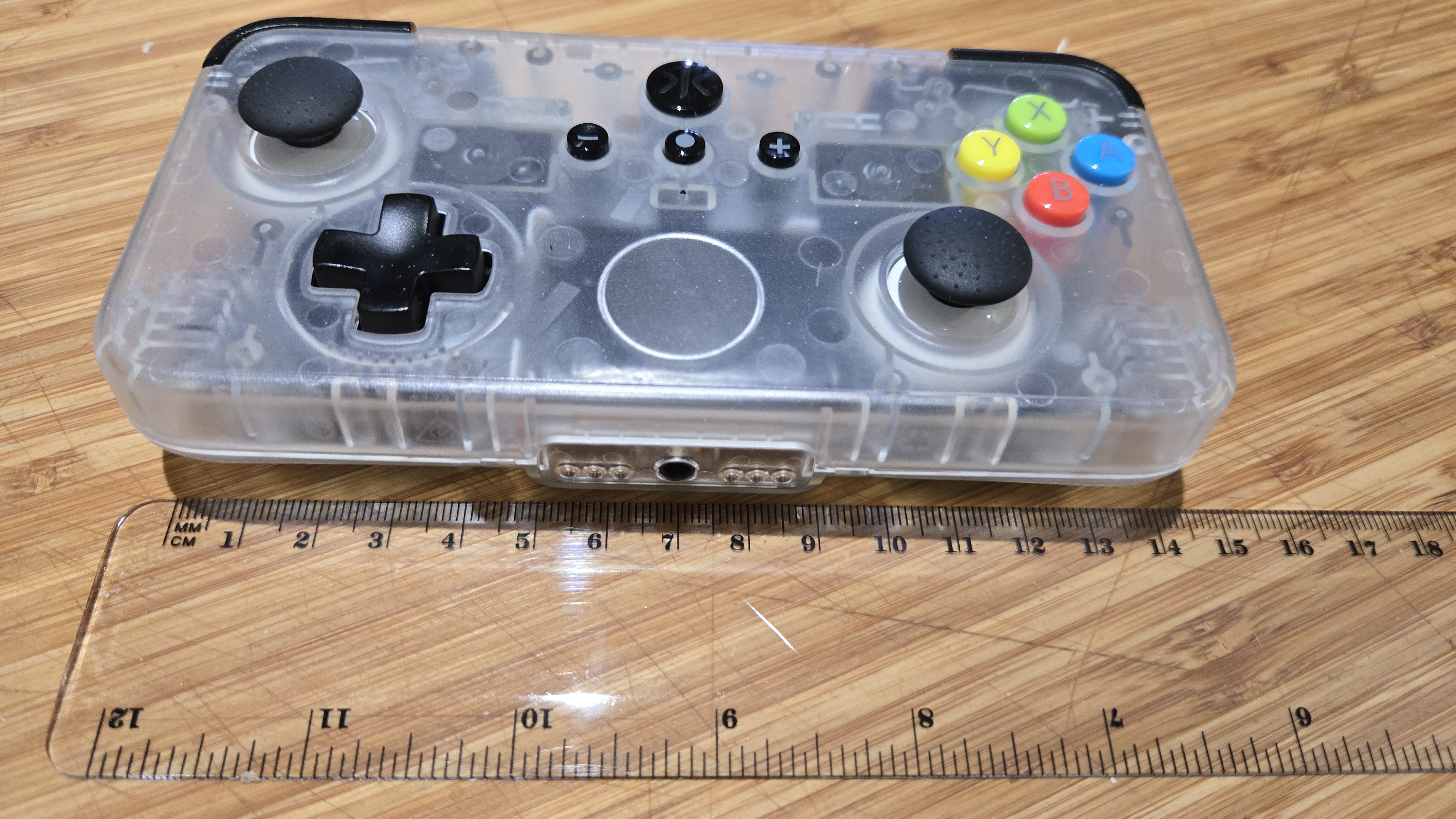 A Classic clear edition of the CRKD NEO S rectangular controller sits on a desk, beside a ruler. The ruler shows that that the compact controller is about 14.5 centimetres in width. The ruler is made out of clear plastic, while the body of the controller is made out of a cloudy clear plastic that allows the viewer to see the internals.