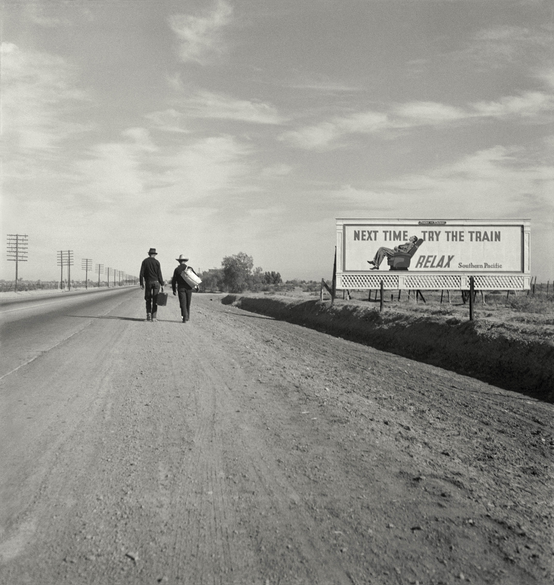 Two men carrying luggage walk away from the camera along a flat California highway, passing a billboard advertising Southern Pacific railroad with the words "Next time try the train &amp;mdash; relax."