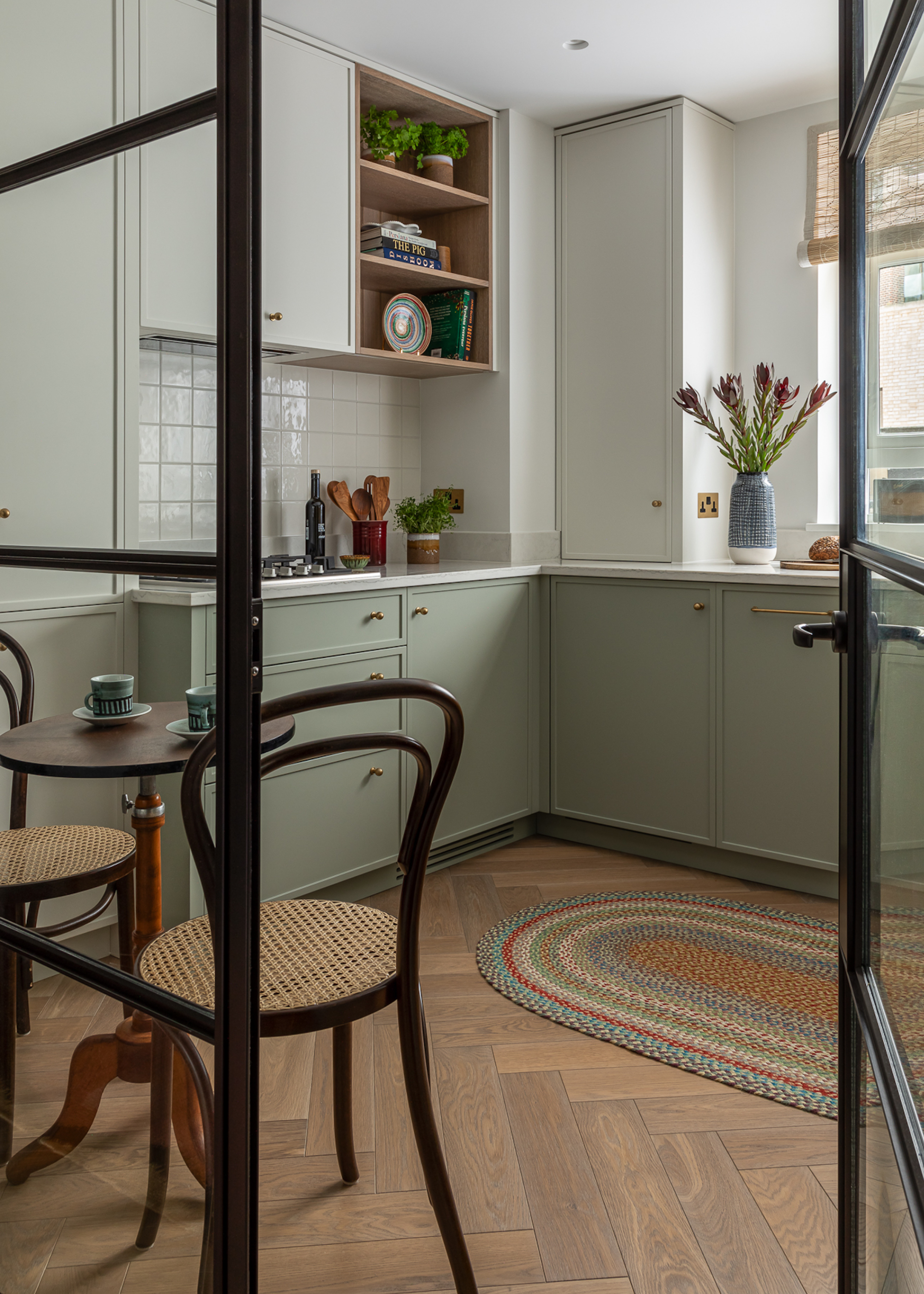 A pale olive colored kitchen with light wood, herringbone flooring. There is a glass divider and door with black detailing, leading through, with a small, wooden table to the left.