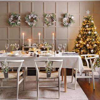 Wishbone style chairs around dining table set for Christmas. Four wreaths on a panelled feature wall and gold lit Christmas tree