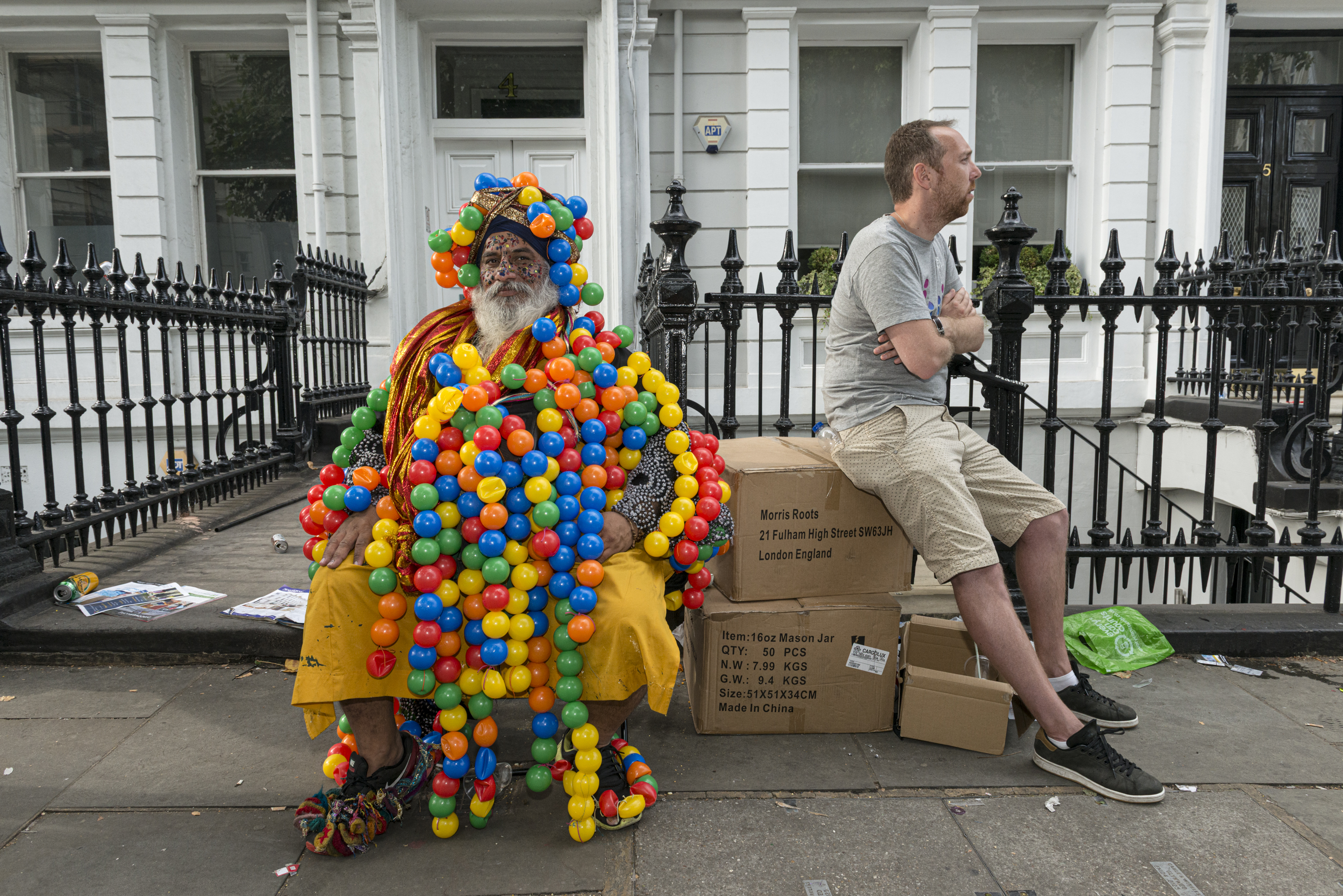 A man sitting on cardboard boxes looks away from a person sitting next to him who is elaborately covered in hundreds of colorful plastic balls.