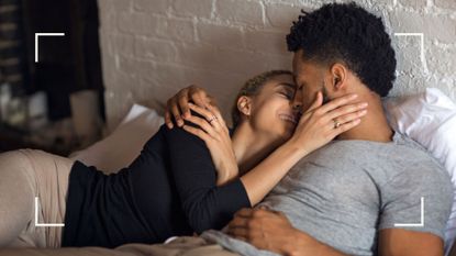 Couple in bed kissing and smiling against white brick painted wall to illustrate the closed missionary sex position