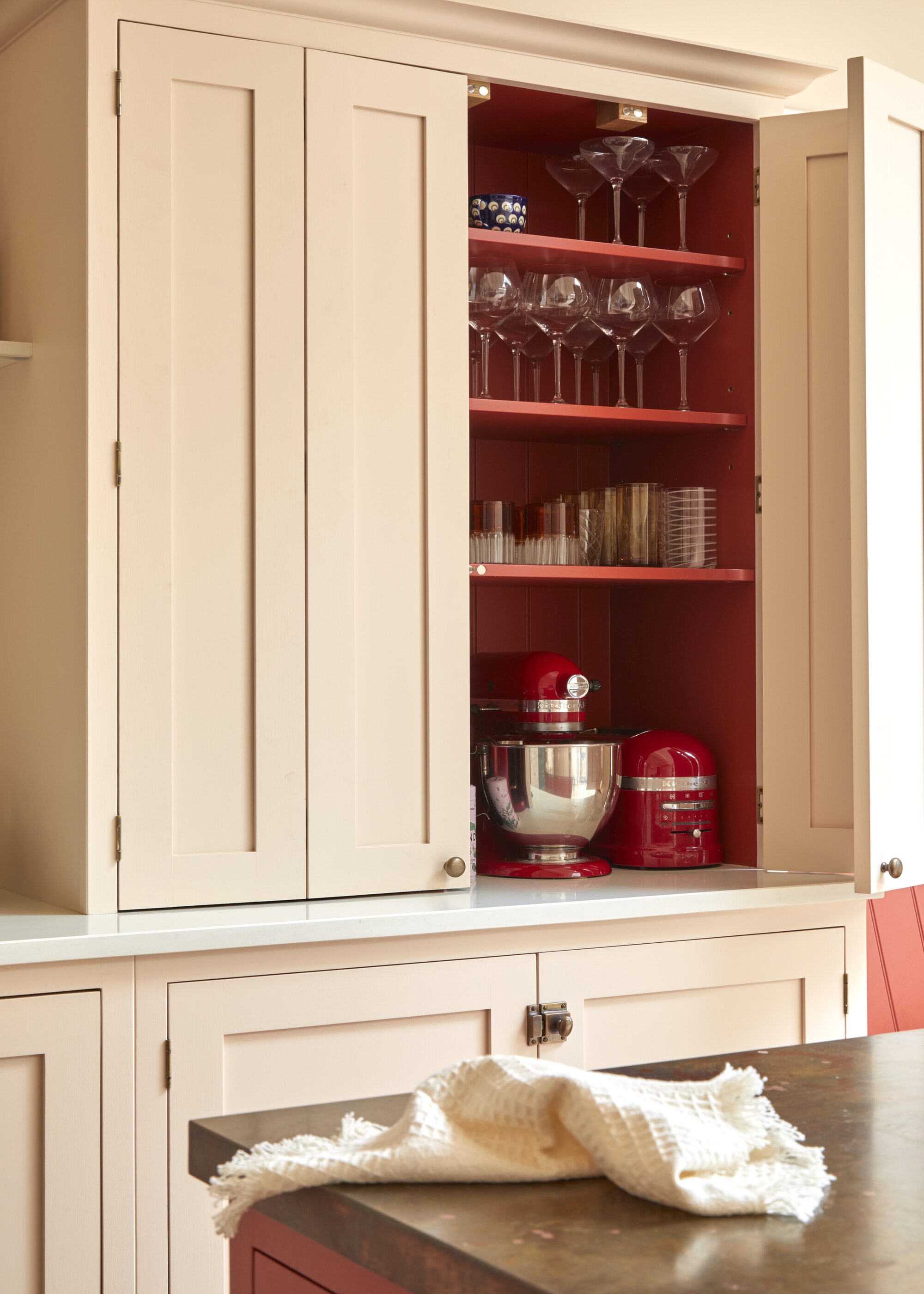 A kitchen pantry cabinet painted in a pale pink color with a red interior and brass latch hardware