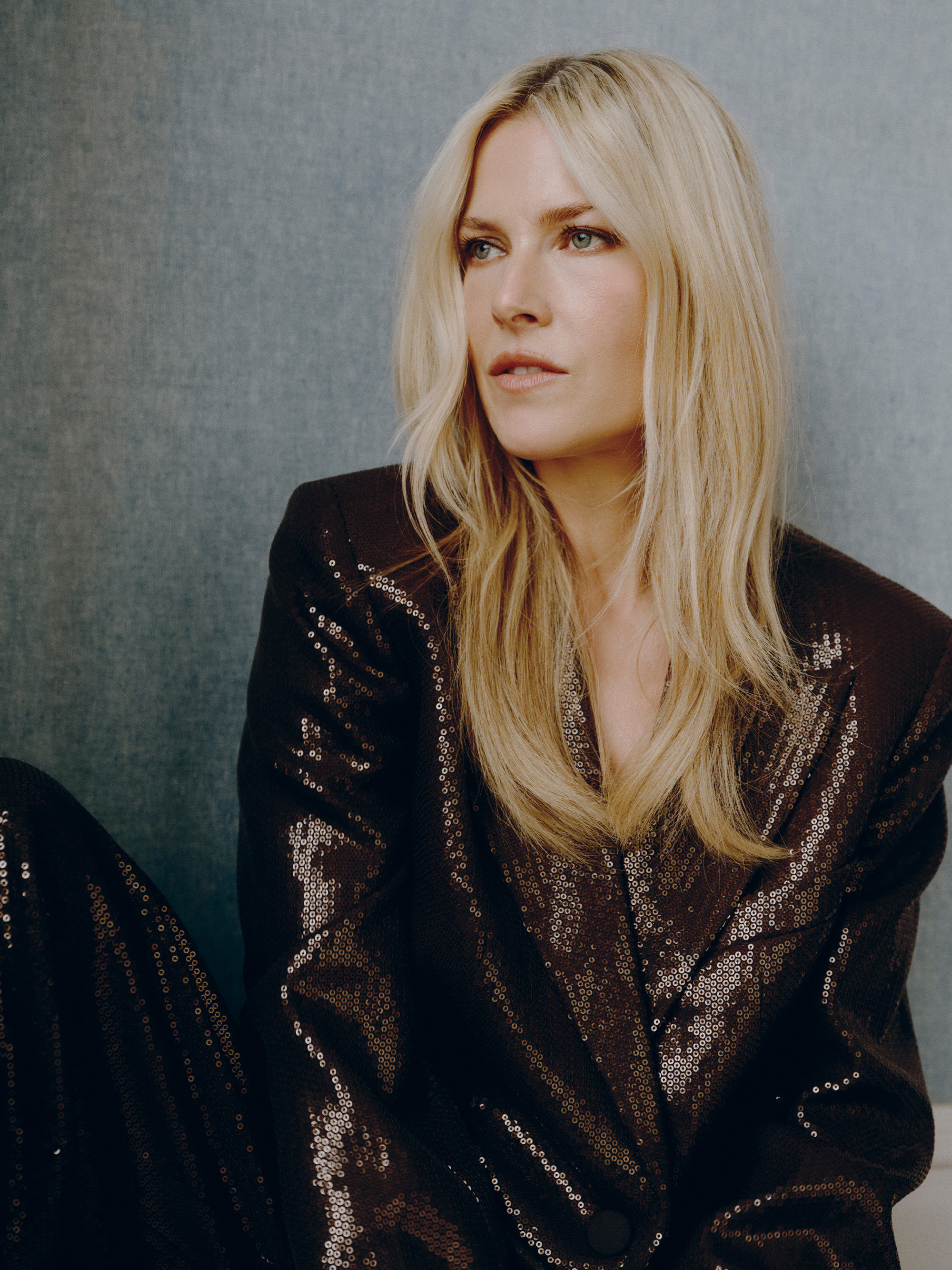Ali Larter sitting in front of a gray background. She is looking off camera and wearing a chocolate brown sequin suit by Michael Kors.
