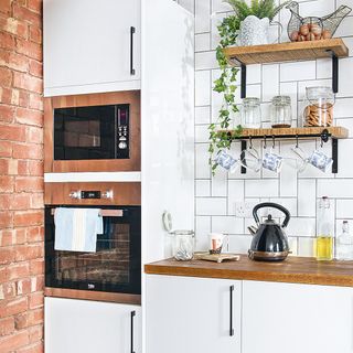 Kitchen with a white tiled and an exposed brick wall, white cabinets, 2 wooden shelves, a wooden countertop