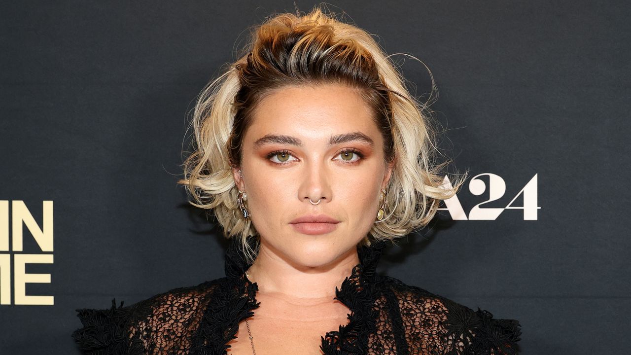 Florence Pugh wearing a black lace top and wavy blond hair standing against a gray background