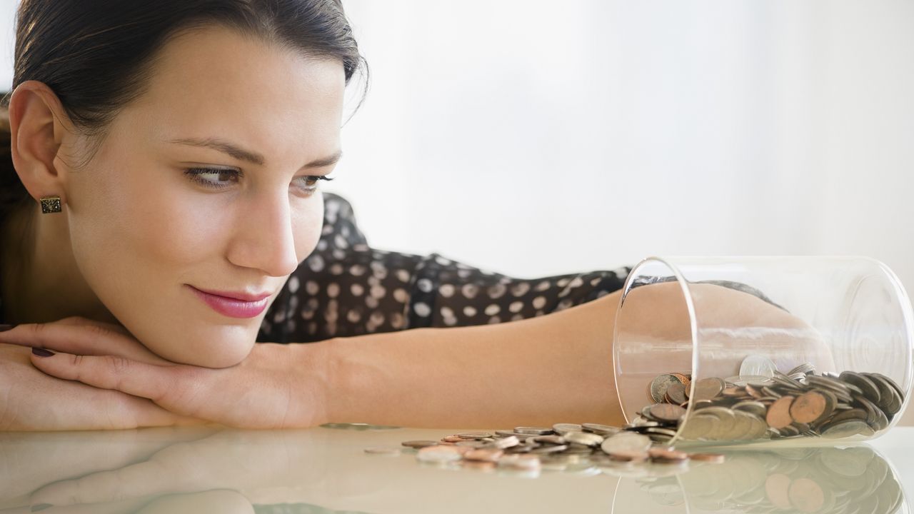 A young woman sits with her chin on her arm, gazing at a tipped-over jar of coins.