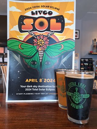 two glasses of beer, one dark and one golden, stand on a wooden table next to a promotional poster featuring a moth and the sun.
