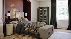 A wooden bed set into a red-painted alcove in a large bedroom. Patterned bedding on the bed and a grey pin-stripe ottoman at the foot. 