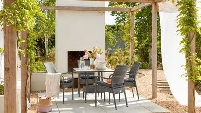 An outdoor living area with an outdoor fireplace, a wooden pergola, and a black outdoor furniture set with four chairs and a square table, and a concrete floor