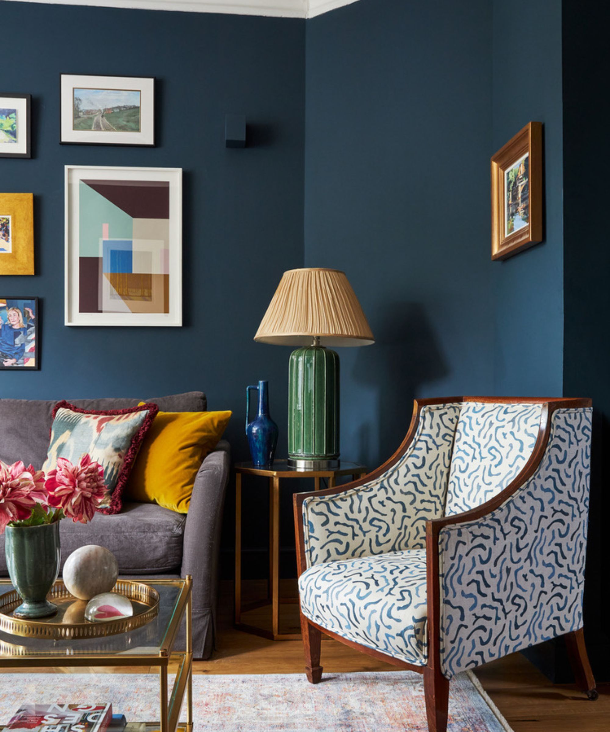 Living room with dark blue walls, gallery wall, upholstered curved antique armchair, grey sofa, glass gold coffee table, and large green table lamp
