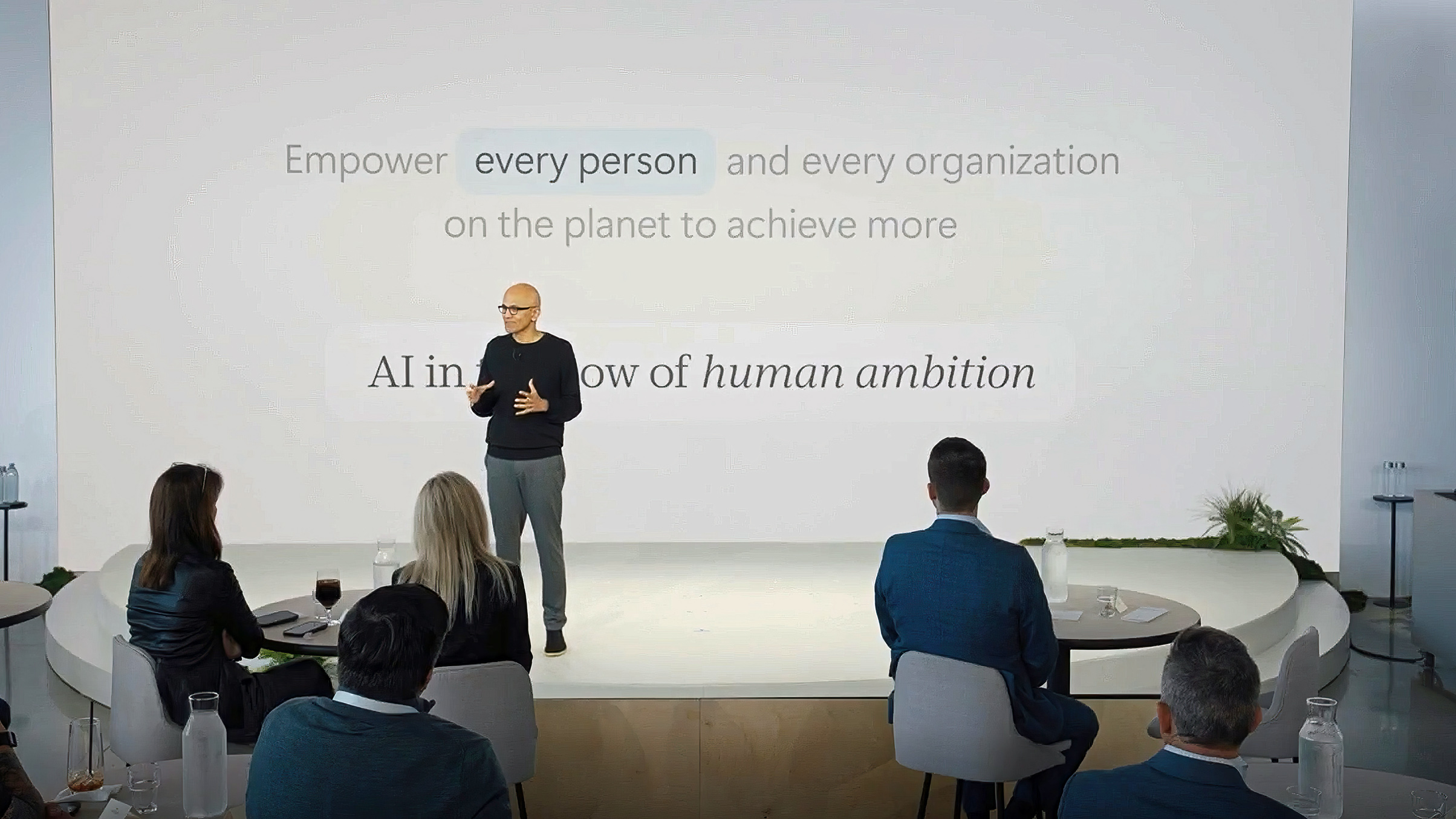 Satya Nadella gestures on stage in front of a white slide reading "Empower every person and every organization on the planet to achieve more," facing an audience.
