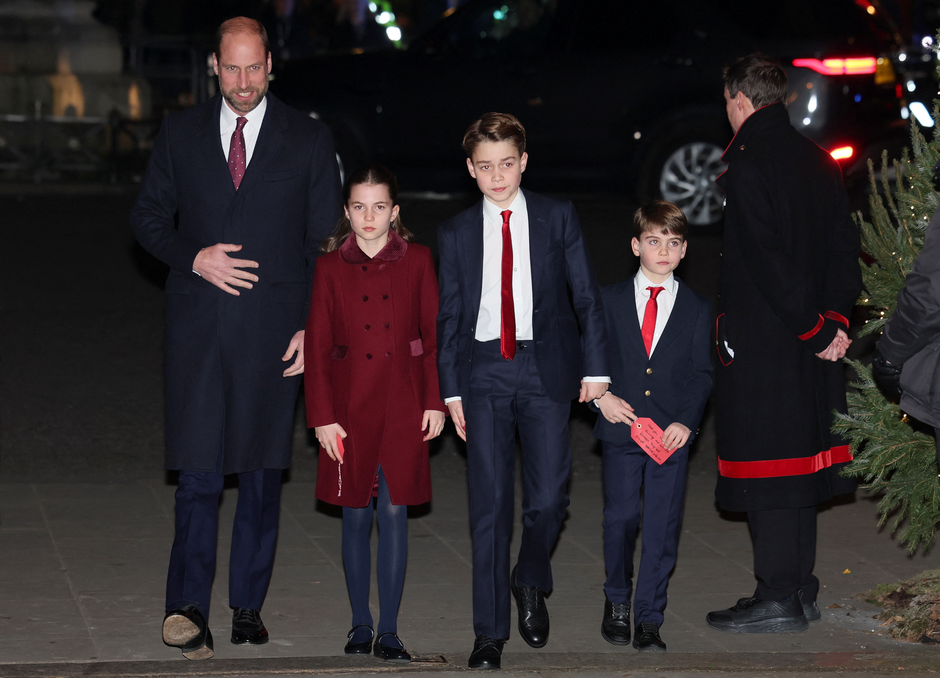 Prince William wears a suit and a long winter coat as he walks beside his three children, Princess Charlotte in a red coat, and sons Prince George and Prince Louis in navy suits with white shirts and red ties