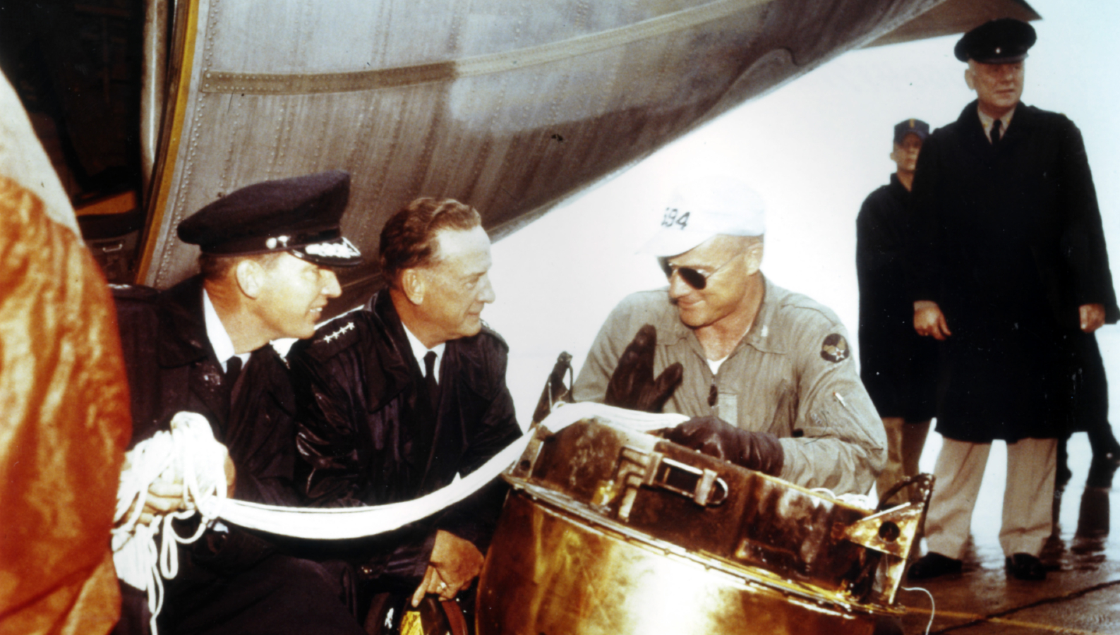 Various men wearing black and tan military uniforms talk to each other leaning over a large metal capsule