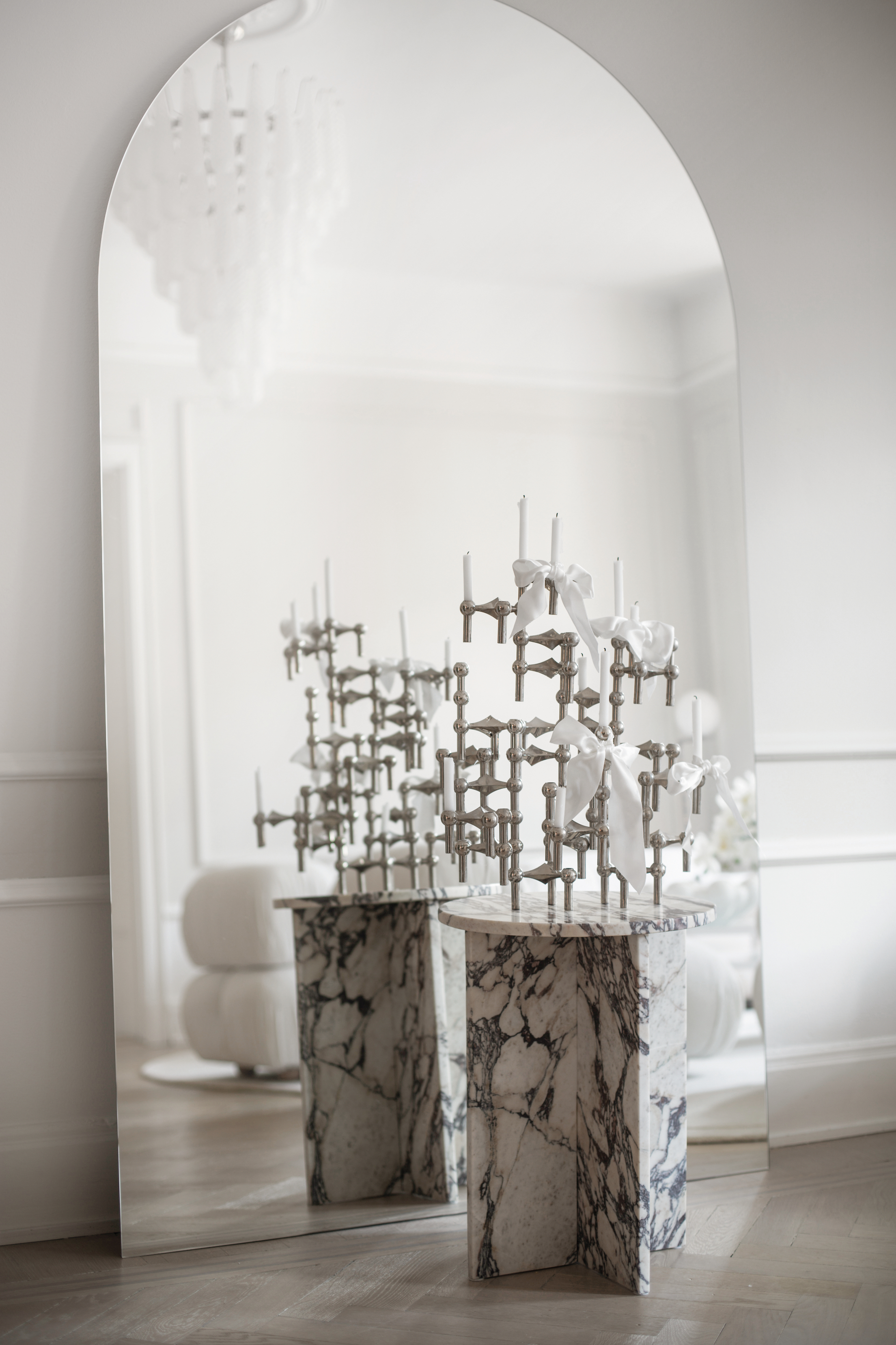 Corner of a living room with a white and grey marble side table and silver candelabra decorated with white bows reflected in a curved floorstanding mirror