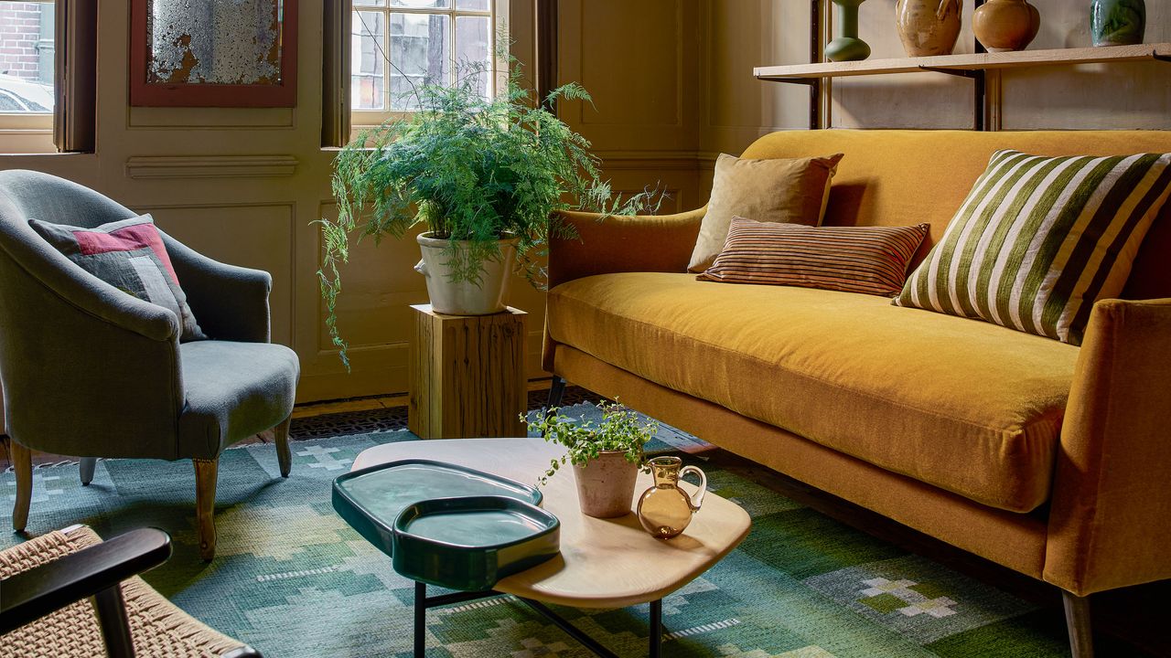 A sunlit, eclectic living room featuring a vibrant mustard yellow velvet sofa adorned with striped and solid earth-toned pillows. To the left, a grey armchair sits on a patterned green and blue area rug. In the center, a light wood coffee table holds dark green ceramic trays, a small potted plant, and a glass pitcher. A large, feathery green fern in a white ceramic pot sits on a wooden pedestal by a window, while a rustic shelf in the background displays various pottery pieces against olive-toned walls.