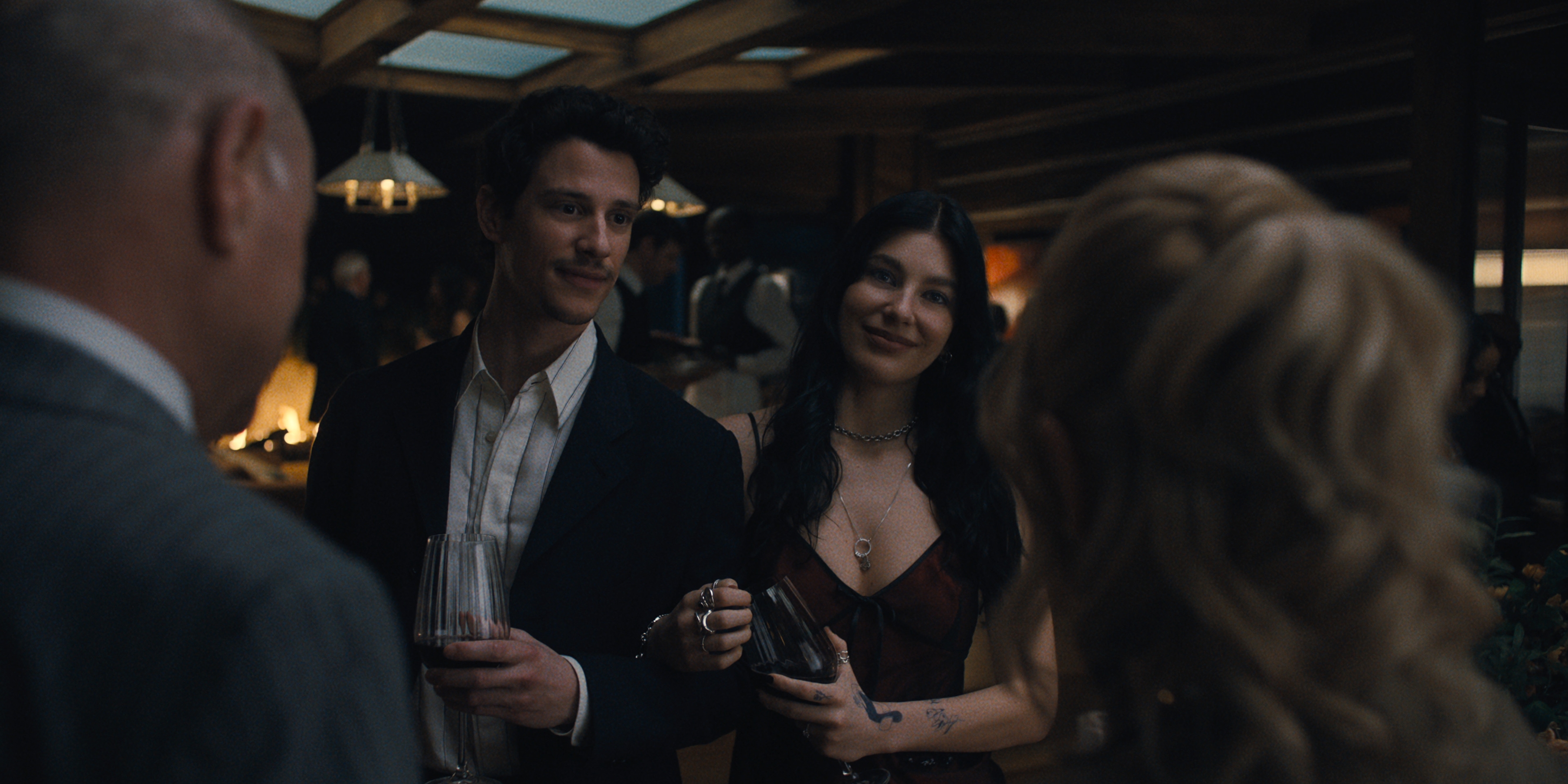 Adam DiMarco as Nicky Cunningham, Camila Morrone as Rachel Harkin in episode 105 of Something Very Bad Is Going To Happen wearing formal wear and standing arm in arm as they talk to another couple at a party