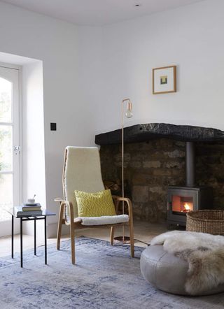 A white room with an old brick fireplace in the wall. There is a white and wooden chair with a long back and a black side table next to it. In front of the chair is a round light gray poof with a fur throw blanket over it