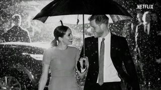 Prince Harry and Meghan MArkle smiling lovingly at each other in the rain under an umbrella in a still from their new Netflix documentary.