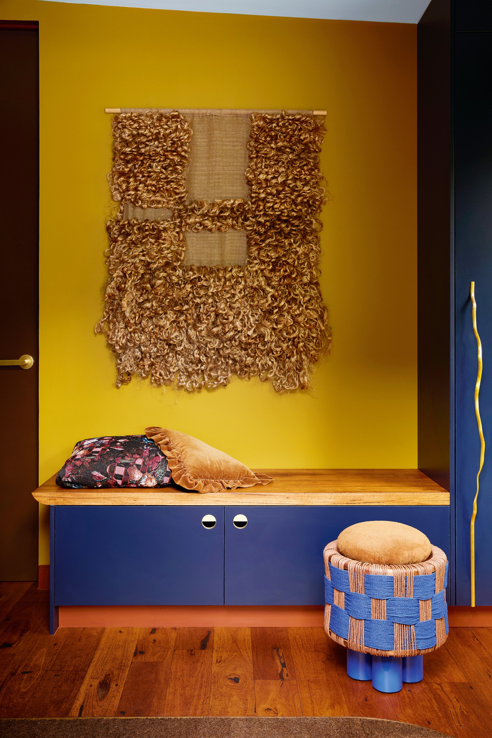 Blue joinery against a yellow wall, with a woven wall hanging above and a blue and tan woven checkerboard footstool