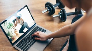 Out-of-focus woman sits on the floor looking at laptop with dumbbells on the floor next to her