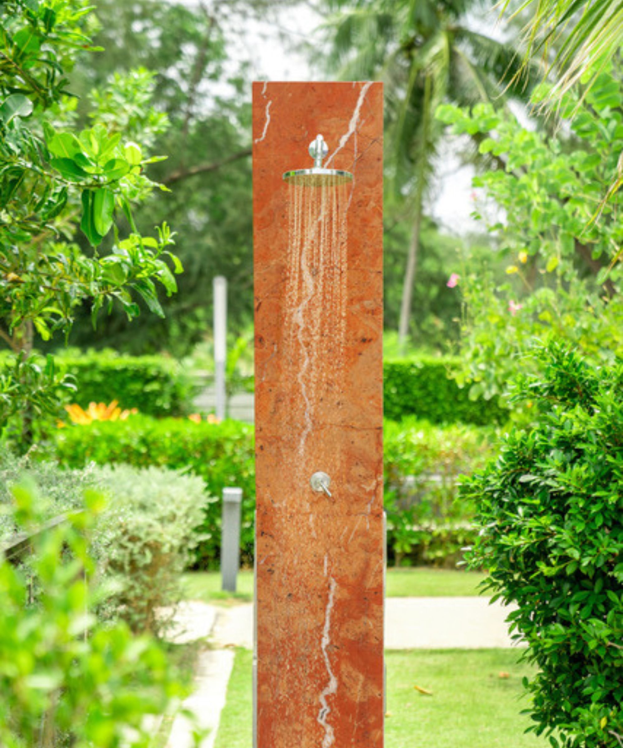A freestanding red marble shower column with a chrome rainfall head running with water, set in a tropical garden with manicured hedging.