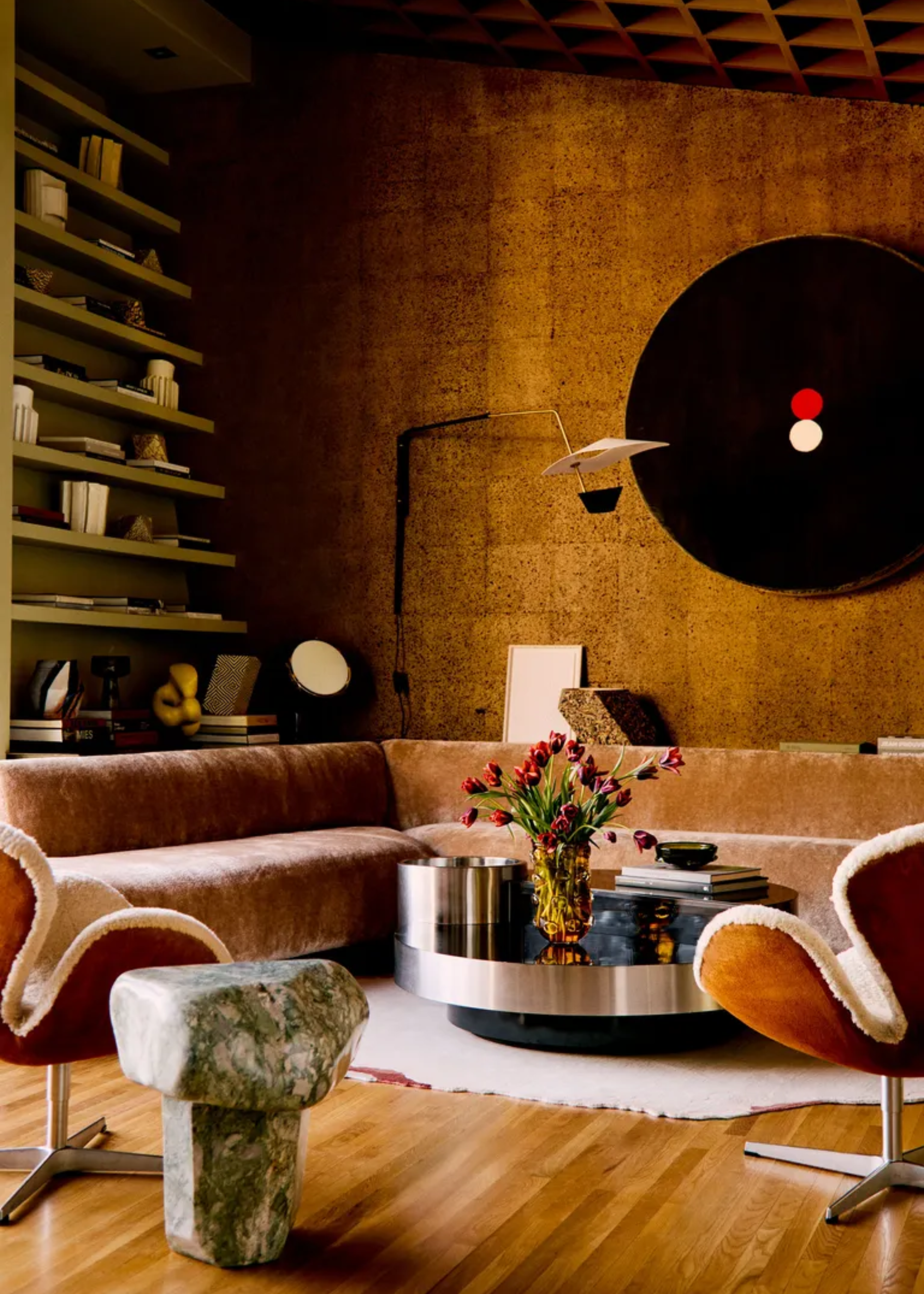 A living room with a cork wall, an open bookshelf, a smooth chenille sofa, a chrome coffee table, and a marble side table