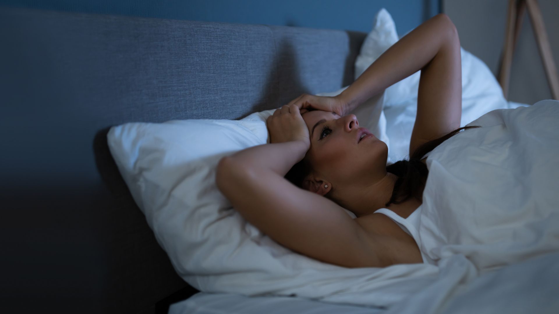 A woman lies awake in bed looking anxious with her hands on her head.