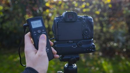 A person hold a shutter release cable on the left with a camera on a tripod to the right. The shutter release is plugged into the camera via a cable.