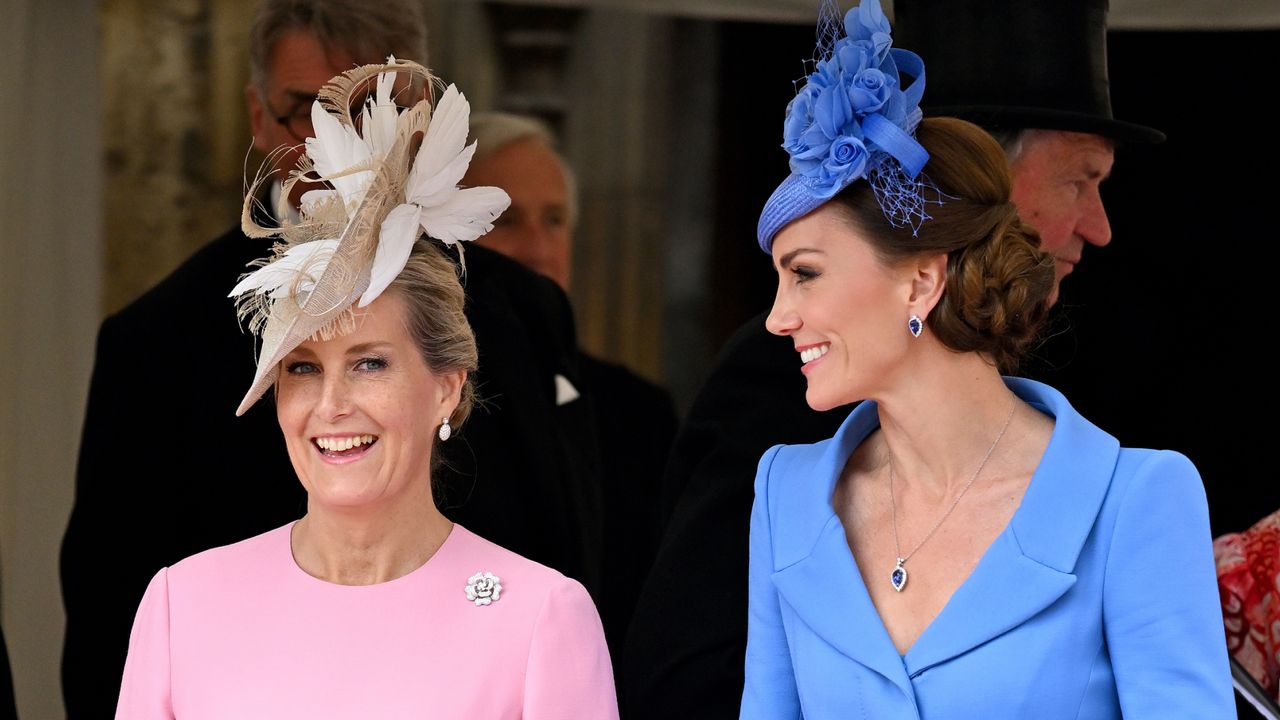 Duchess Sophie wearing a pink dress and feathered tan hat smiling next to Kate Middleton, dressed in a blue long-sleeved coat and matching hat with an updo