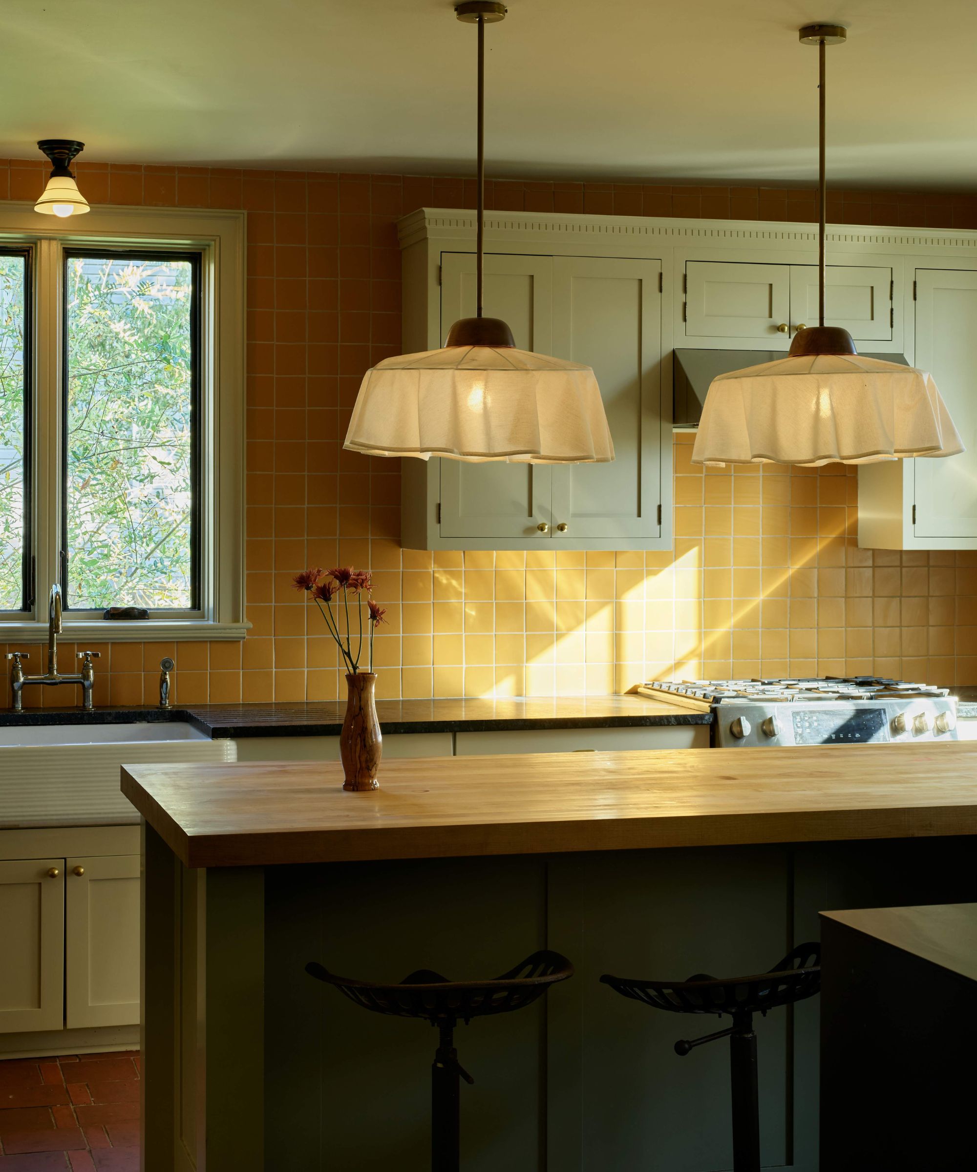 Kitchen with light wood butcher block counter, two white ruffled pendant lights, off-white upper cabinets, and a backsplash made of small, square orange/yellow tiles