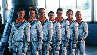 seven men in silver spacesuits stand beside a cone-shaped spacecraft
