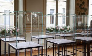 Gallery room, large windows, glass display cabinets with items of jewellery pieces on metal stands inside, floral displays on window sills, wooden floor