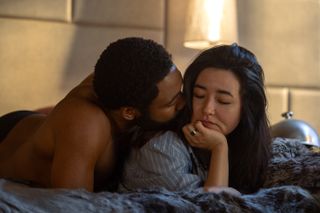 a man (donald glover as john smith) kisses a woman (maya erskine as jane smith) on the cheek as they lay in bed, in 'mr. and mrs. smith'