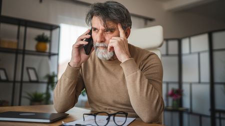 An older man on the phone at his dining room table looks suspicious.