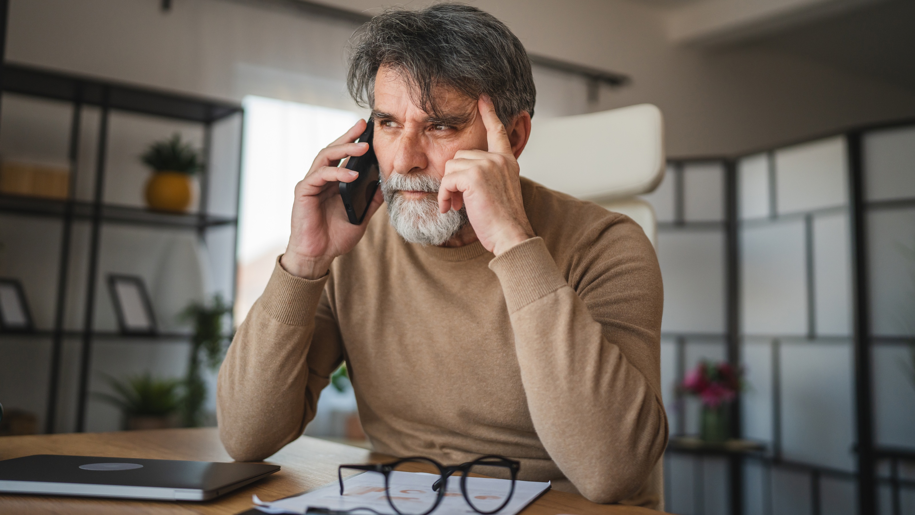 An older man on the phone at his dining room table looks suspicious.