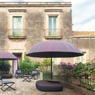 A purple rounded parasol with a sofa under it, chairs and potted plants around it in front of a historical looking two storey house.