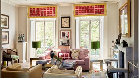 Eclectic living room with large French doors leading out onto a balcony, pink and yellow blinds, pink sofa, coffee table with books and a vase, and two leather armchairs 