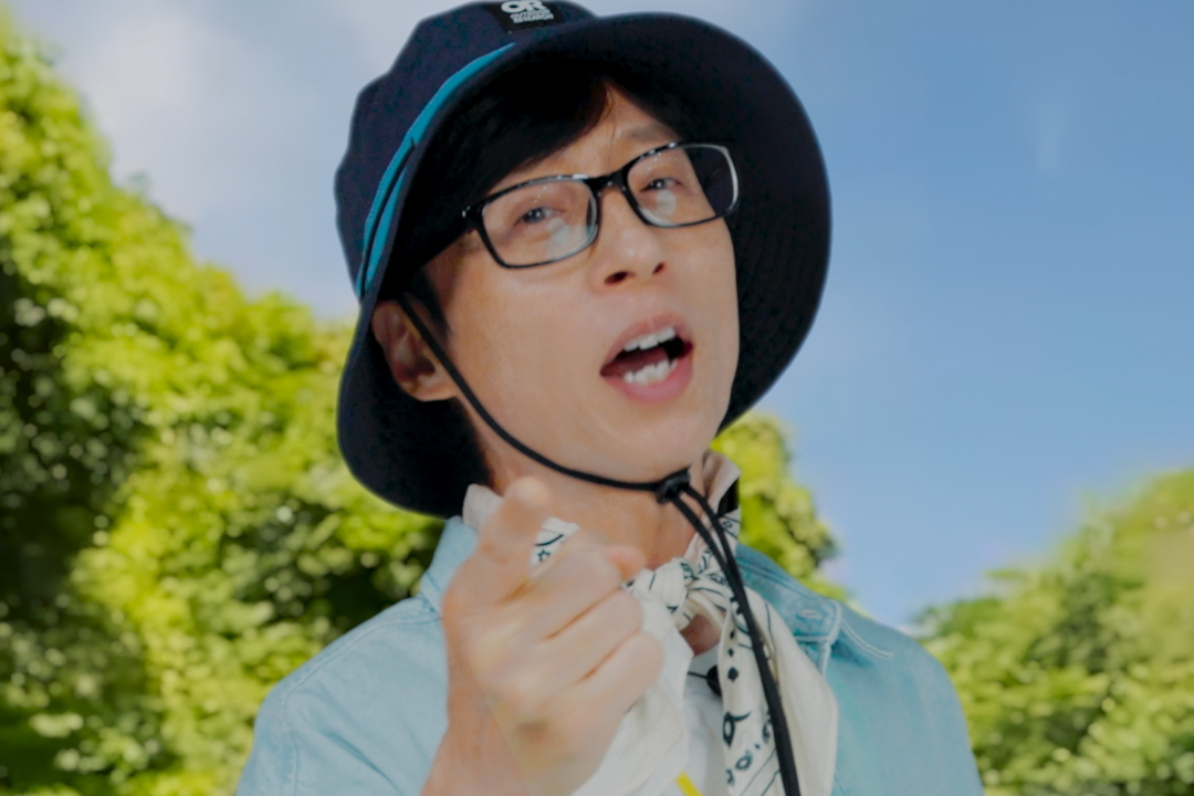 A TV still of Korean entertainer Yu Jae-seok, from the upcoming show 'Jae-seok's B&amp;amp;B Rules!' He wears a bucket hat with a string clinched under his jaw, as he points at the camera while standing outdoors in front of a tree line.