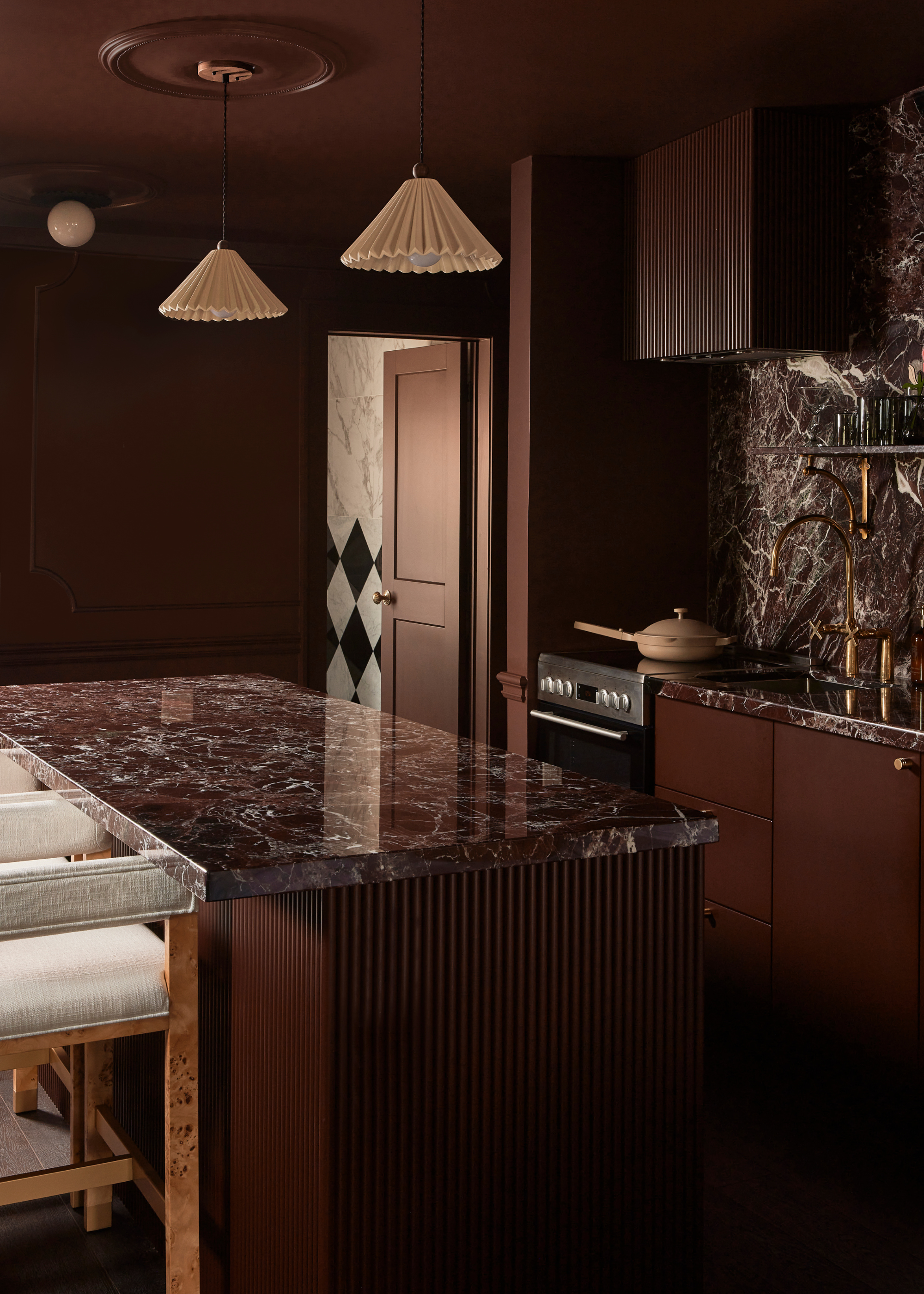 A burgundy drenched kitchen with all sorts of materials and textures including burgundy marble worktop and splashback and white pleated light shades over the island