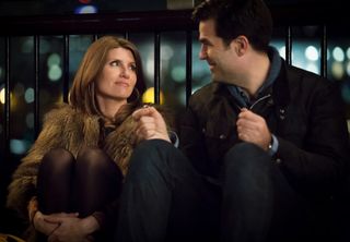 A woman (Sharon Horgan as Sharon Morris) wraps her hands around her legs as she sits next to a man (Rob Delaney as Rob Norris) offering her an engagement ring, in 'Catastrophe.'