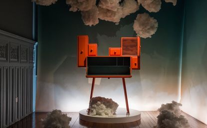 An orange chest of drawers with a stepped design with drawers of different lengths and heights on four thin legs on a stage surrounded by cotton clouds.