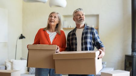 An older couple look around their new home while carrying moving boxes.