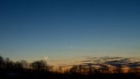 A orange and blue night sky with a glowing crescent moon and a bright dot for the planet Mercury with a series of silhouettes of trees in the foreground
