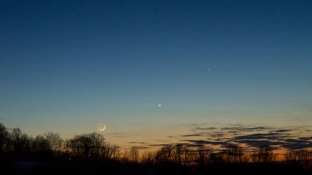 A orange and blue night sky with a glowing crescent moon and a bright dot for the planet Mercury with a series of silhouettes of trees in the foreground