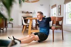 A man performs triceps dips in a living room wearing a t-shirts, shorts, sneakers and sweat bands on his wrists. He is facing away from a chair, with his legs straight out in front of him and both hands resting on the edge of the chair. His elbows point backwards as he's lowering himself down towards the tiled floor. Behind him we see a dining table, fire place, leafy plant and dresser.