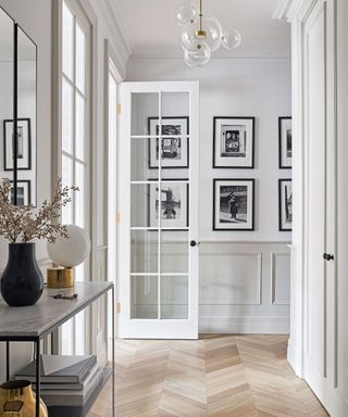 White painted hallway with gray paneling, light wood herringbone flooring, marble console table with opal and brass table lamp, rounded glass geometric pendant, black and white framed pictures, glass paneled doors,
