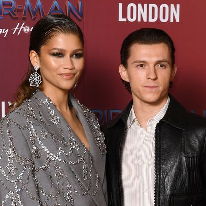 Zendaya wears a silver suit jacket and spider web earrings as she poses with Tom Holland, and Zendaya's small tattoo tribute to Tom Holland is visible in her strapless orange dress